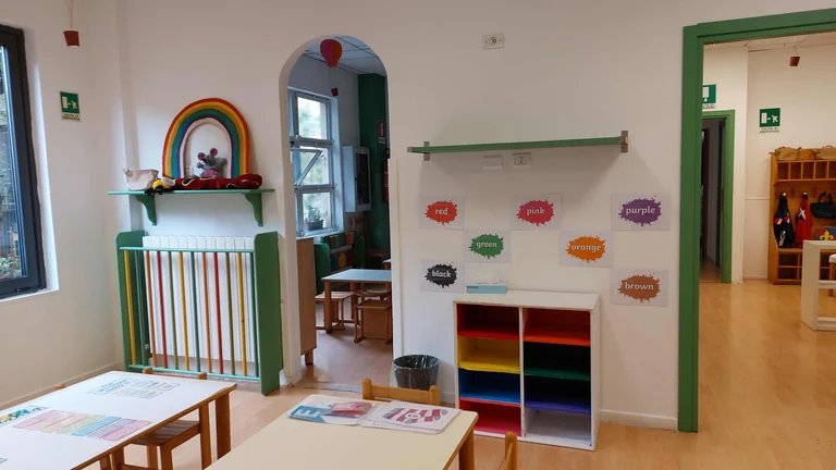 Colorful preschool classroom with wooden tables, rainbow decorations, color-word posters on wall, and shelving with supplies