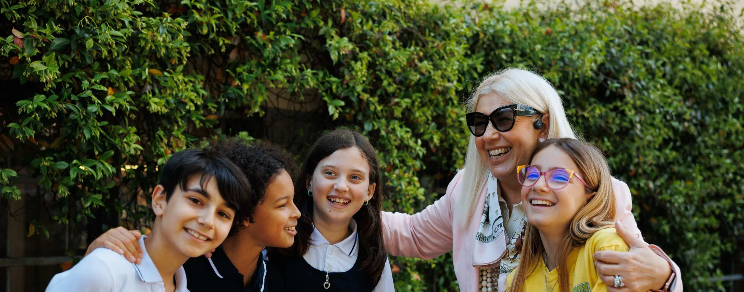 Smiling older woman with sunglasses embracing four diverse school children outdoors against a green hedge backdrop