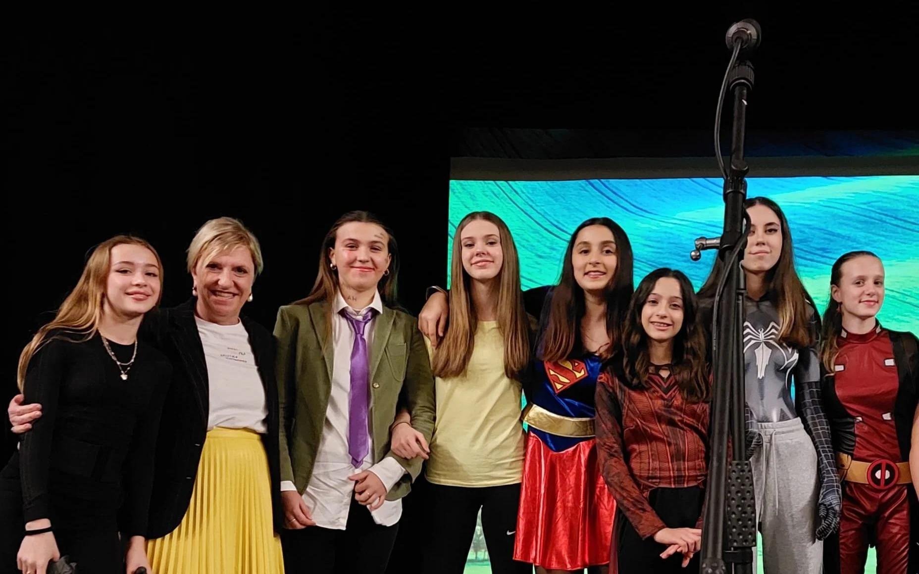 Eight students and a teacher posing on stage in costumes including Supergirl, Spider-Man, and Deadpool at a school event