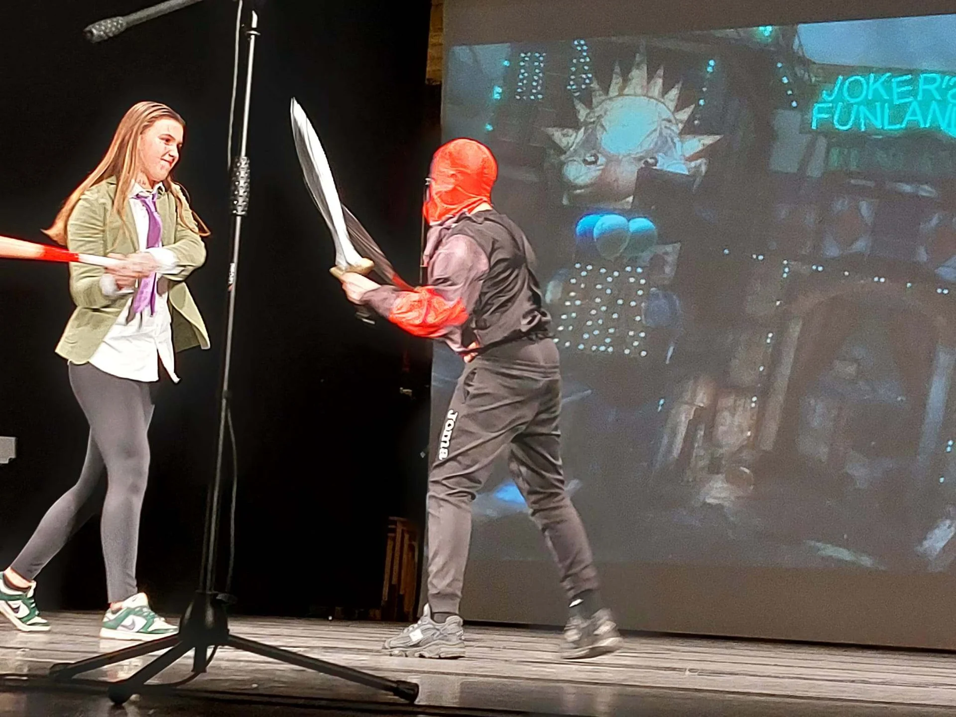 Two students performing on stage with prop weapons, one wearing a red mask, with a 