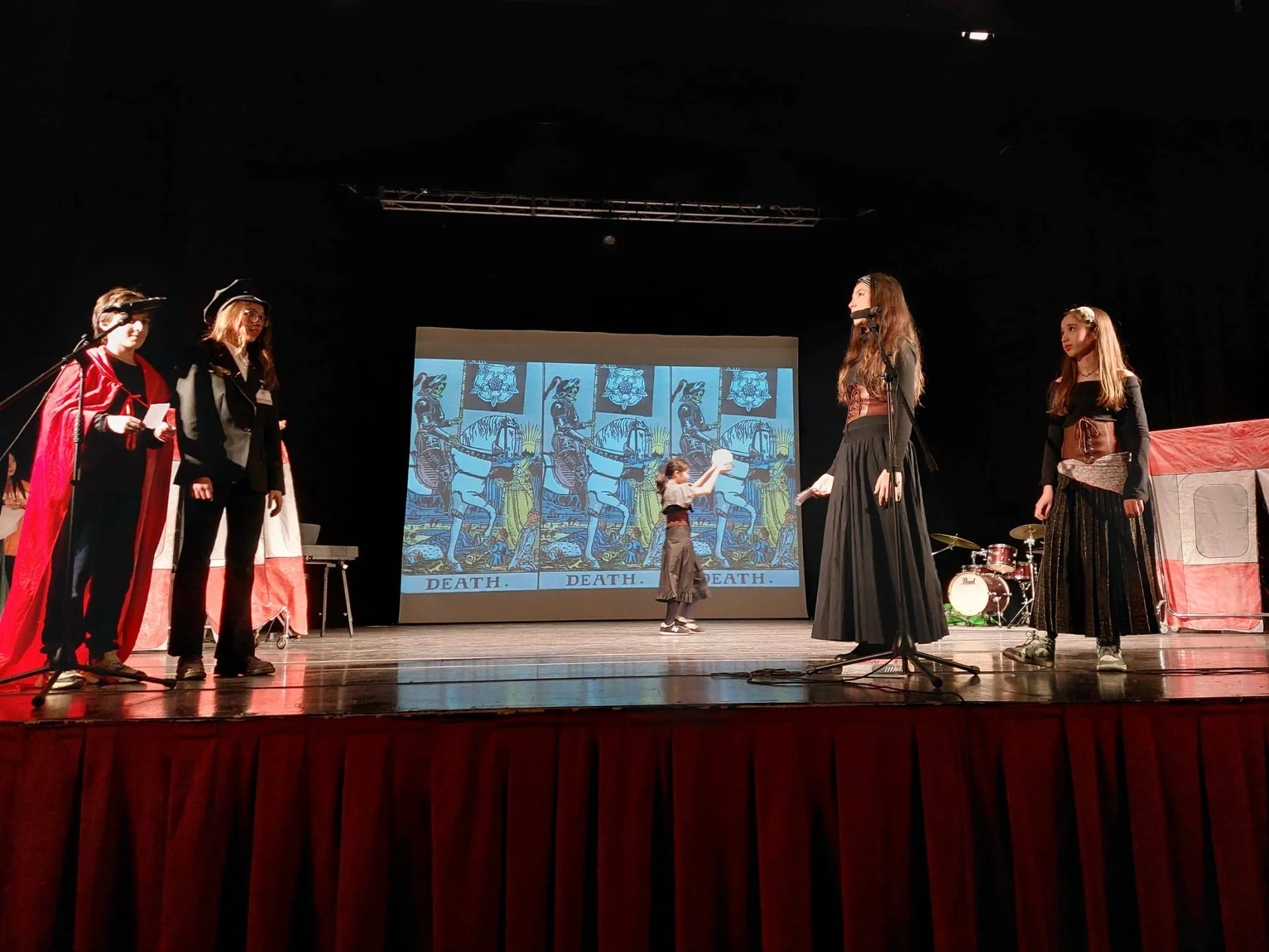 Students performing a theatrical production on stage with medieval costumes, tarot card backdrop displaying 