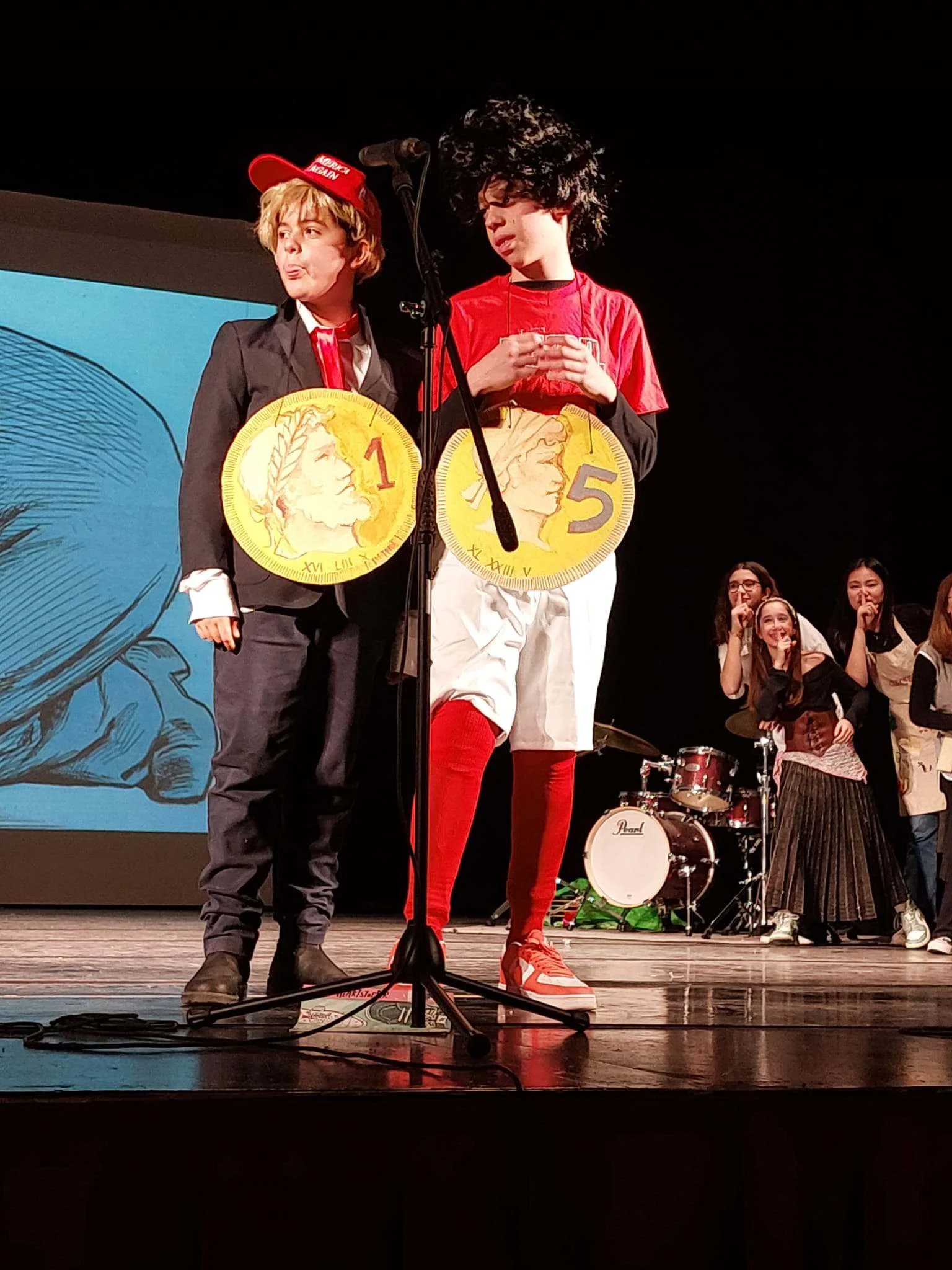 Two students on stage wearing large gold coin props numbered 1 and 5, performing in a school theatrical production with classmates watching